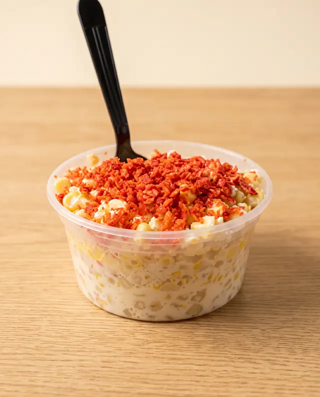 Un vaso de esquites, elote y queso cotija bajo una capa de chips picantes triturados