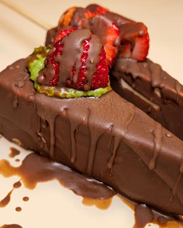Una paleta de helado cubierta de chocolate escurriendo, con una fresa con chocolate y crema de pistache
