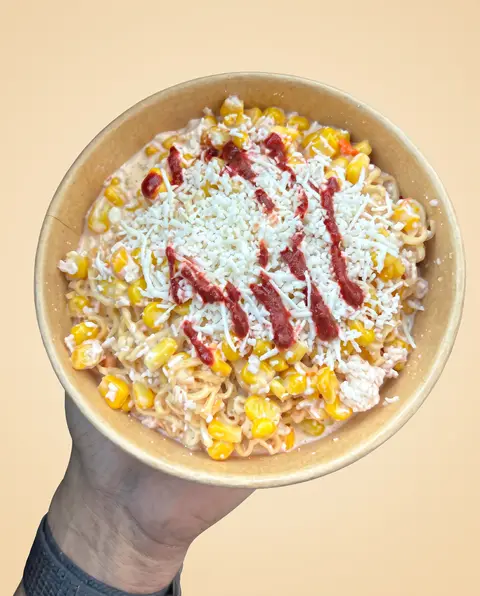 Una mano sosteniendo un vaso de chaska Maruchan: ramen y elote con queso cotija y chamoy