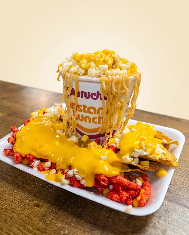 Queso amarillo derretido cayendo sobre un vaso Maruchan con elote y chips picantes