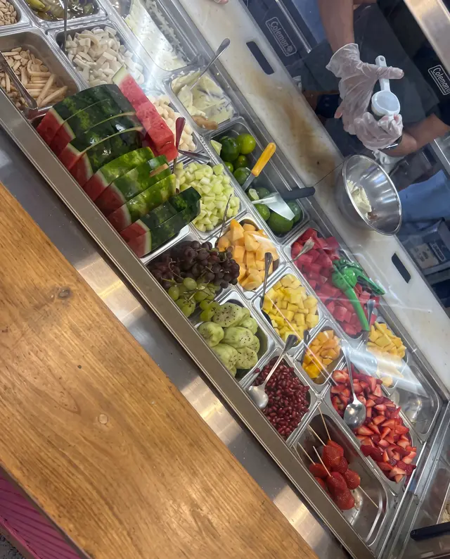 La barra de toppings de Poloco: sandía, pepino, uvas, piña, granada y fresa en compartimentos de acero, con un empleado sirviendo