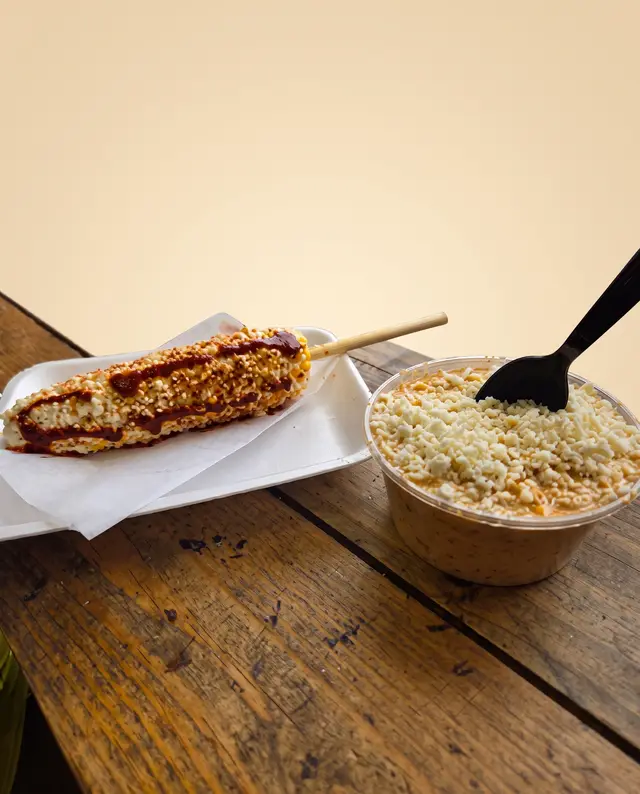 A whole elote on a stick coated in mayo, cheese and chili, next to a cup of esquites