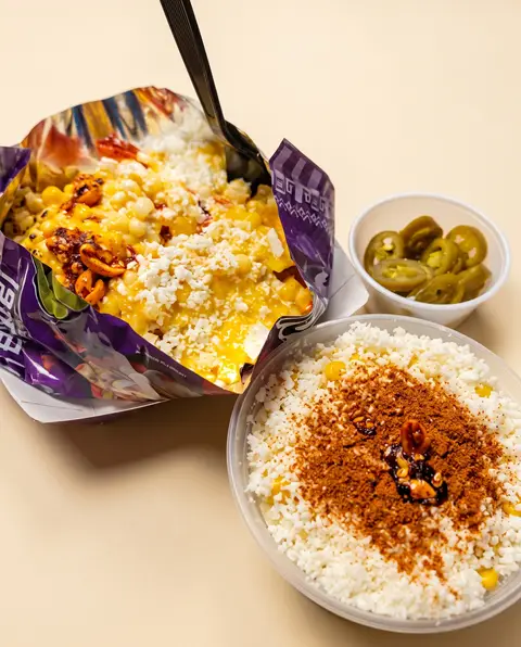 Una bolsa de chips cargada de elote y queso cotija, con jalapeños y un vaso de esquites