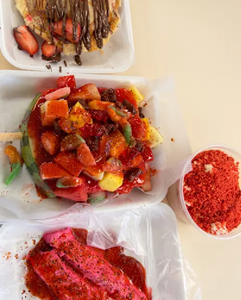 Una charola de fresa y mango con chamoy y gusanitos de goma, junto a un vaso de chamoy en polvo