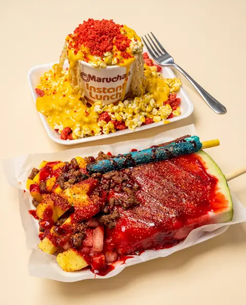 Un vaso Maruchan sobre esquites junto a una rebanada de sandía con chamoy, mango, cueritos y un popote de dulce