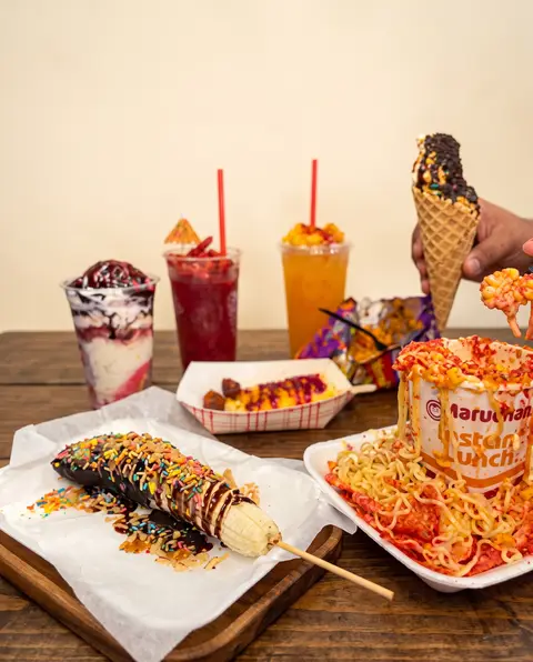 Una mesa llena de Poloco: parfaits, dos aguas frescas, un choco-banana con chispas en palito, un vaso Maruchan con chips picantes y una mano con un cono de waffle