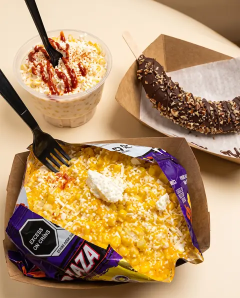 Vista aérea de un vaso de esquites con chamoy, un plátano con chocolate y nuez, y una bolsa de chips cargada
