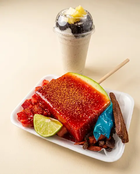 Una rebanada de sandía con chamoy, limón y cueritos junto a una malteada de cookies and cream